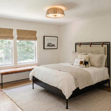 A transitional bedroom with the jute and aged brass Pascal Flush Mount 18" installed in the ceiling center above a black bed frame and leather bench.
