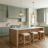 A transitional kitchen features two aged brass and smoke taupe glass globe pendants over a white marble island with sage green cabinetry and wood slatted details.
