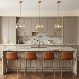 A contemporary kitchen with three Arianne Medium Pendants in aged brass and smoke taupe glass over a white marble waterfall island with taupe cabinets and leather stools.