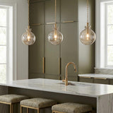 A contemporary kitchen featuring three Arianne aged brass and smoke taupe glass globe pendants suspended over a white marble waterfall island and olive green cabinets.