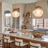 A transitional kitchen with two two-tone art glass and brass pendant lights hanging over a marble island with light wood barstools and beige cabinets.