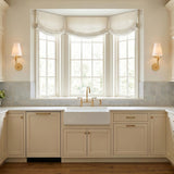 A transitional kitchen with two Junia Wall Sconce 17", Textured Gold fixtures with linen shades flanking a large bay window above a white farmhouse sink and cream cabinets.