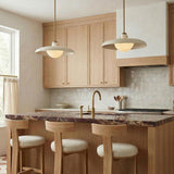 A contemporary kitchen features two Bain Large Pendant 20", Off-White and Patina Brass fixtures over a dark marble island with light wood cabinets and a tile backsplash.