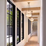 A modern farmhouse foyer with two patina brass and milk glass pendants spaced along the ceiling between wood beams, featuring large black-framed windows and light wood floors.