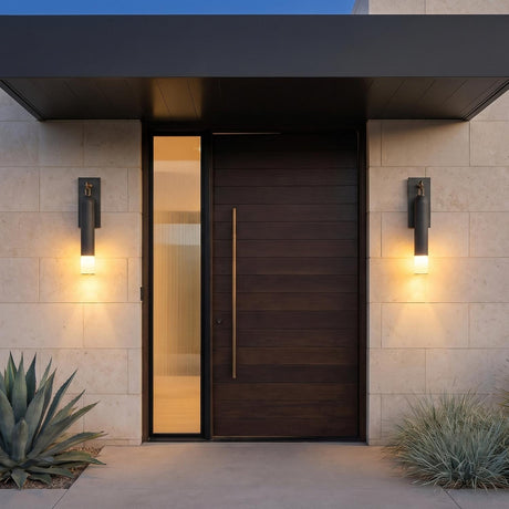 A modern home entry with two graphite and patina brass Langley Outdoor Wall sconces flanking a dark wood door on beige stone walls with agave landscaping at dusk.