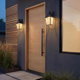 A modern entryway with Baelor Outdoor Wall Light lanterns in textured black with patina brass accents flanking a light wood door set against dark stucco and wood slats.
