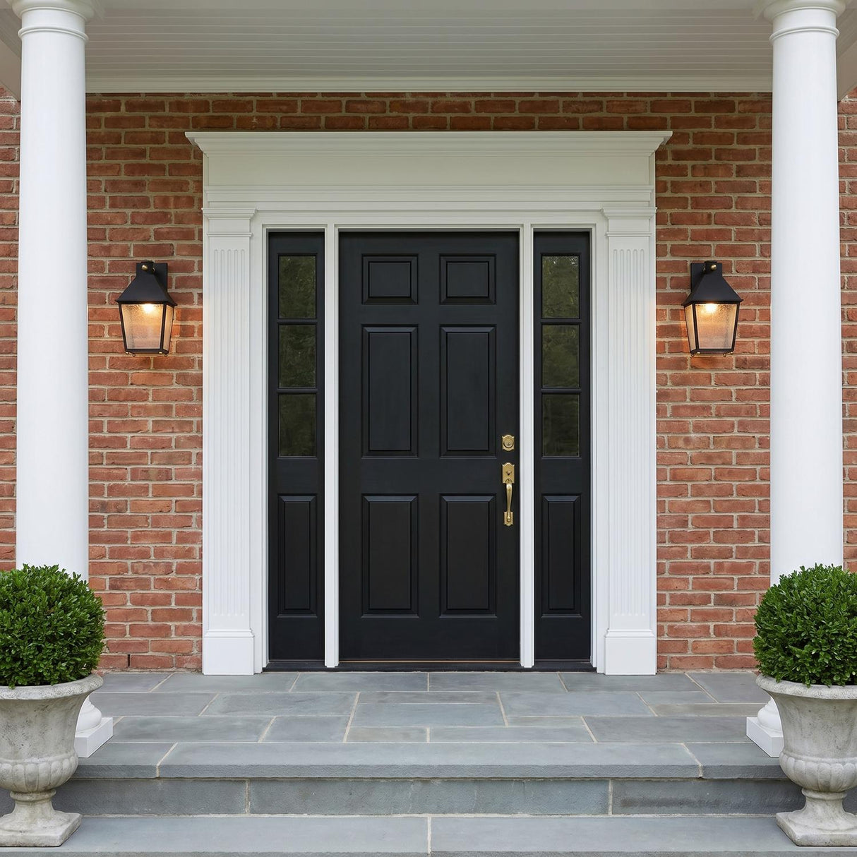 A Colonial red brick entrance with two Baelor Outdoor Wall Lights in textured black flanking a black front door at dusk featuring boxwood shrubs and stone steps.