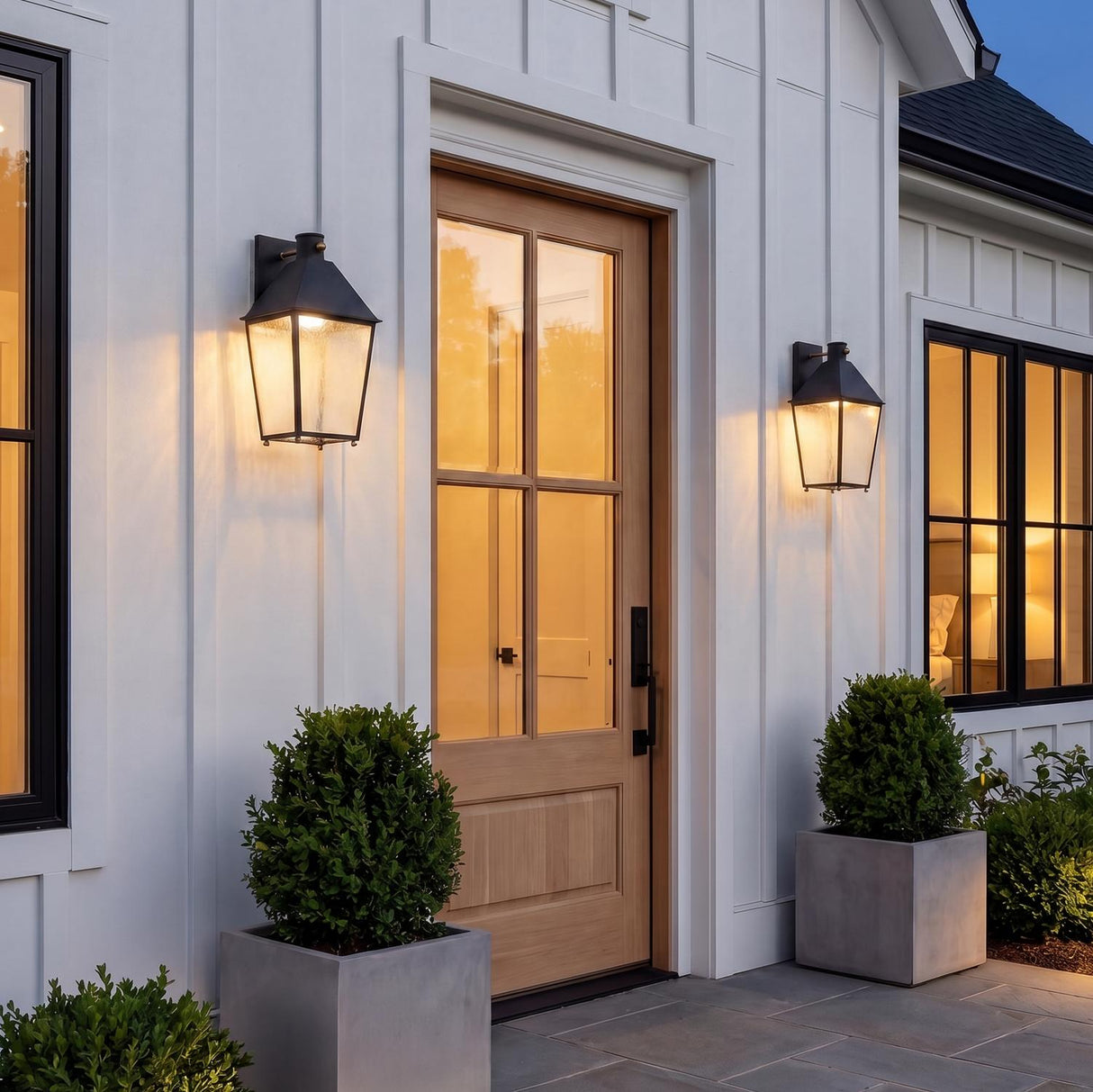 A modern farmhouse entryway featuring two textured black lanterns with patina brass accents flanking a light wood front door on white board and batten siding.