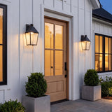 A modern farmhouse entryway featuring two textured black lanterns with patina brass accents flanking a light wood front door on white board and batten siding.