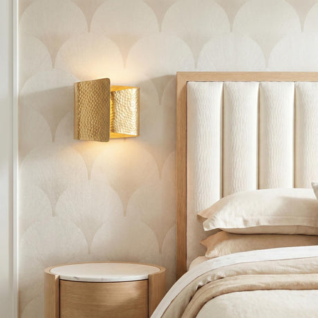 A transitional bedroom featuring a hammered aged brass Trey LED Wall Sconce 8 inch mounted on fan-patterned wallpaper beside a channel-tufted headboard and oak nightstand.