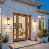 A modern farmhouse entry featuring patina brass wall lanterns flanking a light wood door on white horizontal siding with potted palm plants at twilight.