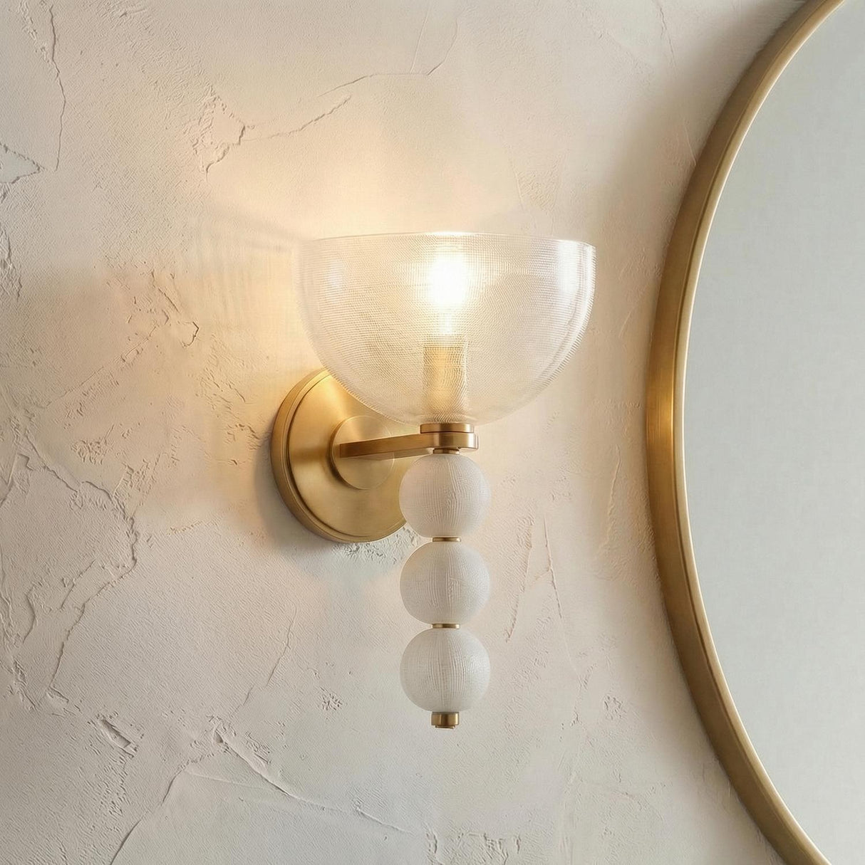 A contemporary bathroom featuring an Amity Wall Sconce in aged brass and textured glass installed on a plaster wall beside a round gold-rimmed mirror.