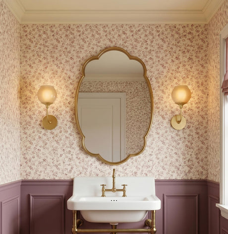A traditional bathroom with the Danica Wall Sconce 12", Aged Brass and Champagne Glass, flanking a scalloped gold mirror above mauve wainscoting and a floral wallpapered wall.