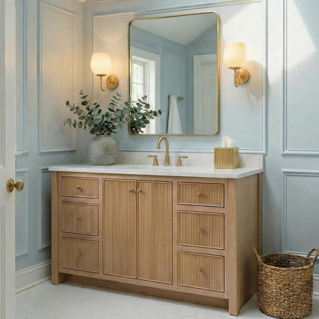 A transitional bathroom with two Goldie wall sconces in aged brass and buttermilk glass flanking a gold mirror above a fluted wood vanity with white quartz.