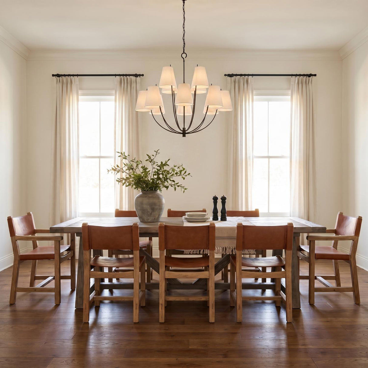 A transitional dining room featuring the Cordelia 12 Light Chandelier 40", Aged Iron with linen shades centered over a wood table and cognac leather chairs.