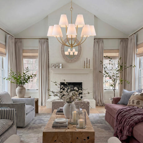 A transitional living room with the Cordelia 12 Light Chandelier 40", Modern Brass featuring white linen shades suspended from a vaulted ceiling above a white brick fireplace.