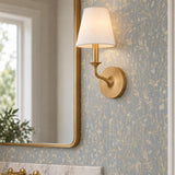 A transitional bathroom with the Harwick Wall Sconce in gilded brass and a white fabric shade mounted beside a mirror on blue floral wallpaper.