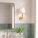 A transitional bathroom features a gilded brass wall sconce with a white fabric shade mounted beside a brass mirror above seafoam green ceramic tile.