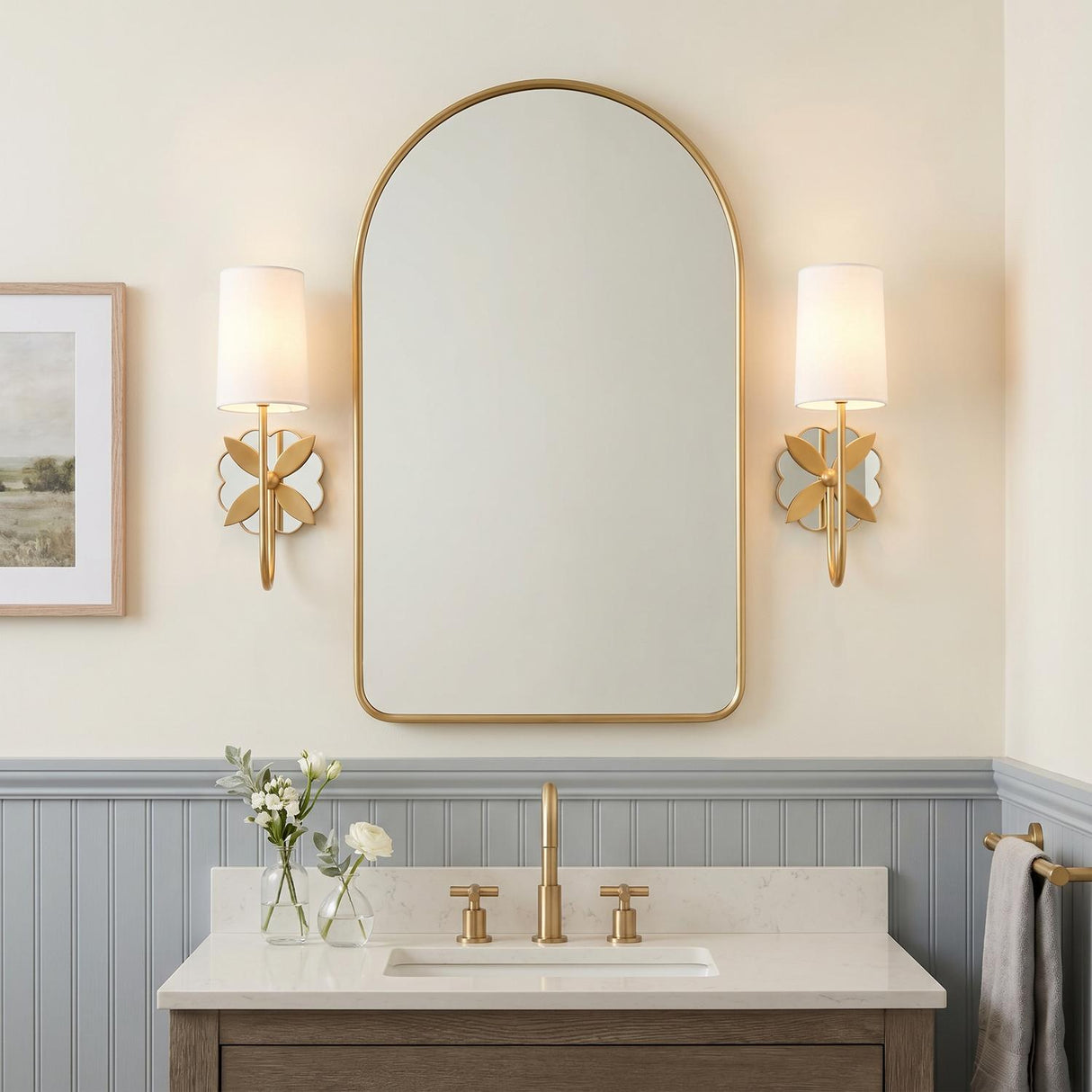A transitional bathroom featuring two Ashby Wall Sconces in modern brass flanking an arched mirror above a white quartz vanity and blue-gray beadboard wainscoting.