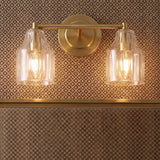 A traditional bathroom features the Radwell 2 Light Vanity 13", Gilded Brass mounted over a mirror against a brown patterned wallpaper with clear ribbed glass shades.