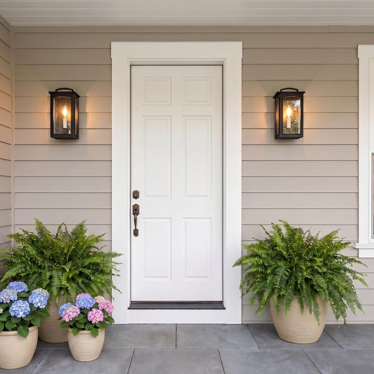 A modern farmhouse entry with two Barlow Outdoor Wall Lights in textured black flanking a white front door on tan siding with ferns and hydrangeas at twilight.