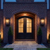An outdoor entryway featuring two Barlow textured black lanterns flanking dark arched double doors on a red brick facade with square stone pavers.