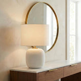 A contemporary entryway featuring a matte white ribbed ceramic table lamp with a linen shade on a marble top walnut console before a gold-rimmed mirror.