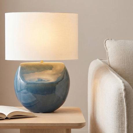 A contemporary living room featuring the Tivola Table Lamp 27", Glossy Cerulean with a white linen shade on a light wood table next to an open book.