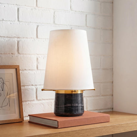 A modern interior featuring the Branson Rechargeable Table Lamp with a white linen shade and aged brass accents placed on a wooden shelf against a white brick wall.
