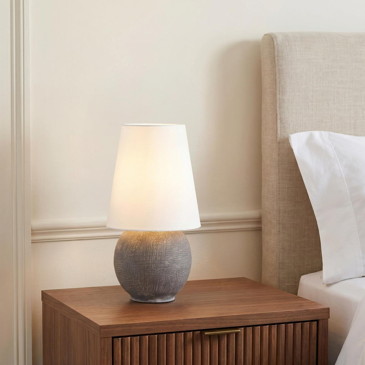 A contemporary bedroom featuring the Inara Rechargeable Table Lamp 15", Artisan Ceramic Gray with a white linen shade on a fluted walnut nightstand beside a beige headboard.