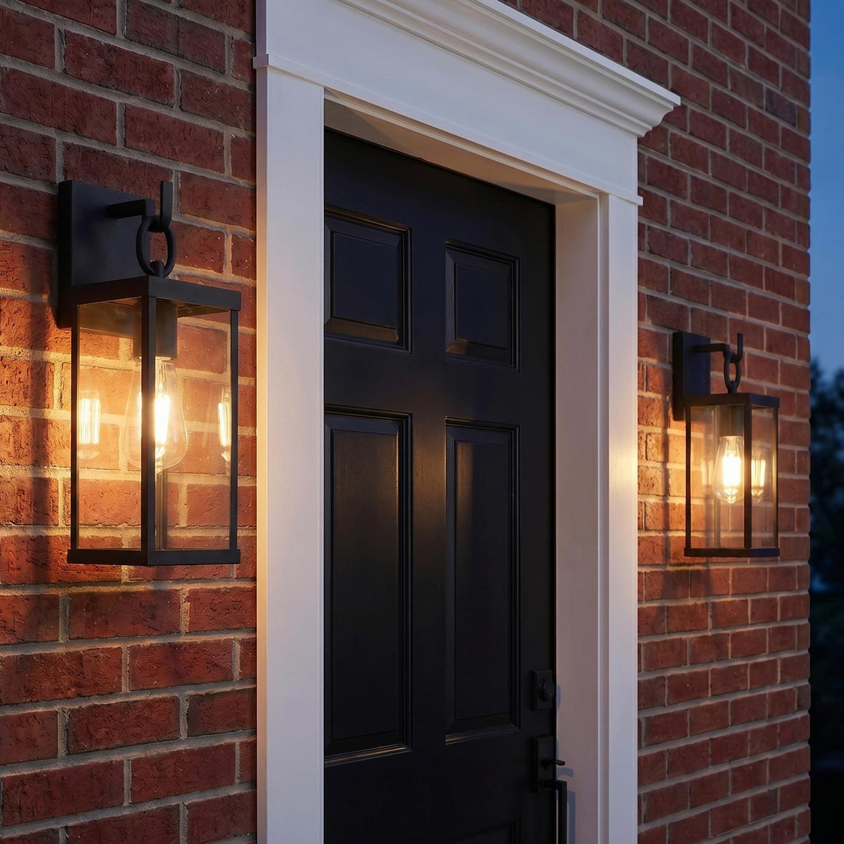 A traditional home exterior with two Lavelle Outdoor Wall Light, Small 15", Textured Black lanterns flanking a black paneled door on a red brick wall at twilight.