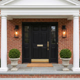 A Colonial red brick entry with two glowing Lavelle Outdoor Wall Light fixtures in textured black flanking a black door with white columns and boxwood topiaries.