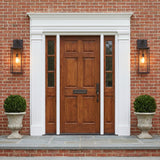 A Colonial red brick entry with two old bronze Lavelle Outdoor Wall Light lanterns flanking a wood front door at dusk with boxwood topiaries in stone urns.