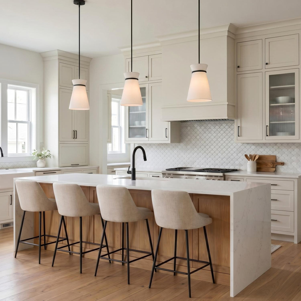 A transitional kitchen with three matte black and white linen pendants over a white marble waterfall island featuring wood accents and cream shaker cabinets.