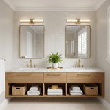 A contemporary bathroom with two satin brass Wendell LED Vanity Lights mounted above rectangular gold mirrors over a floating oak vanity with white marble countertops and woven baskets.