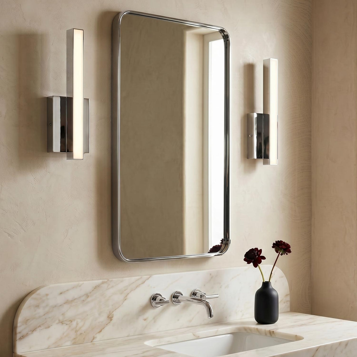 A modern bathroom with two polished chrome Garrison LED Wall Sconce 13", Chrome fixtures flanking a rounded rectangular mirror over a marble vanity and integrated backsplash.
