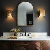A contemporary bathroom featuring two Egan LED Wall Sconces in vintage brass flanking an arched mirror over a white marble countertop and chevron wood vanity.