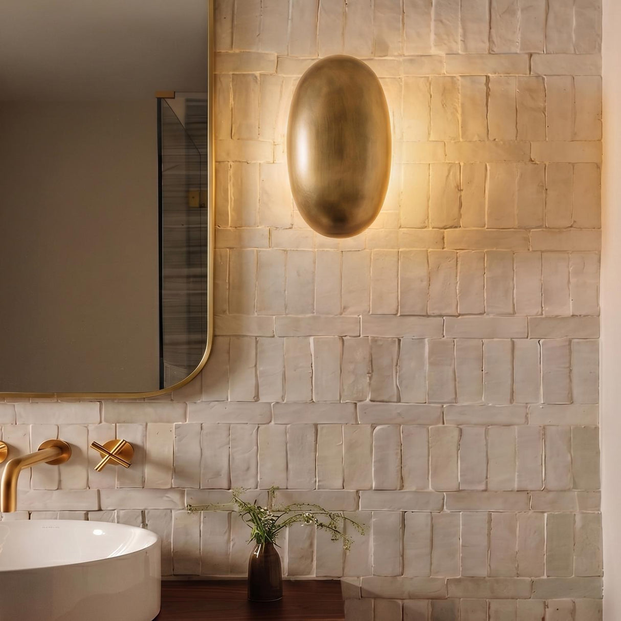 A modern bathroom with a vintage brass Albie LED Wall Sconce 10 inch mounted on a textured white tile wall beside a rounded mirror and brass faucet.