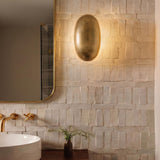 A modern bathroom with a vintage brass Albie LED Wall Sconce 10 inch mounted on a textured white tile wall beside a rounded mirror and brass faucet.