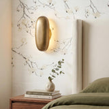 A transitional bedroom with the Albie LED Wall Sconce in vintage brass mounted on a floral mural wall beside a cream wingback headboard and wood nightstand.