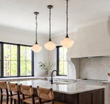 A modern farmhouse kitchen featuring three matte black pendants with vintage milk glass shades over a white marble island with dark wood cabinets and woven bar stools.