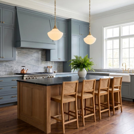 A transitional kitchen with two brushed brass Reva Pendants featuring vintage milk glass shades over a natural wood island with black counters and blue cabinets.