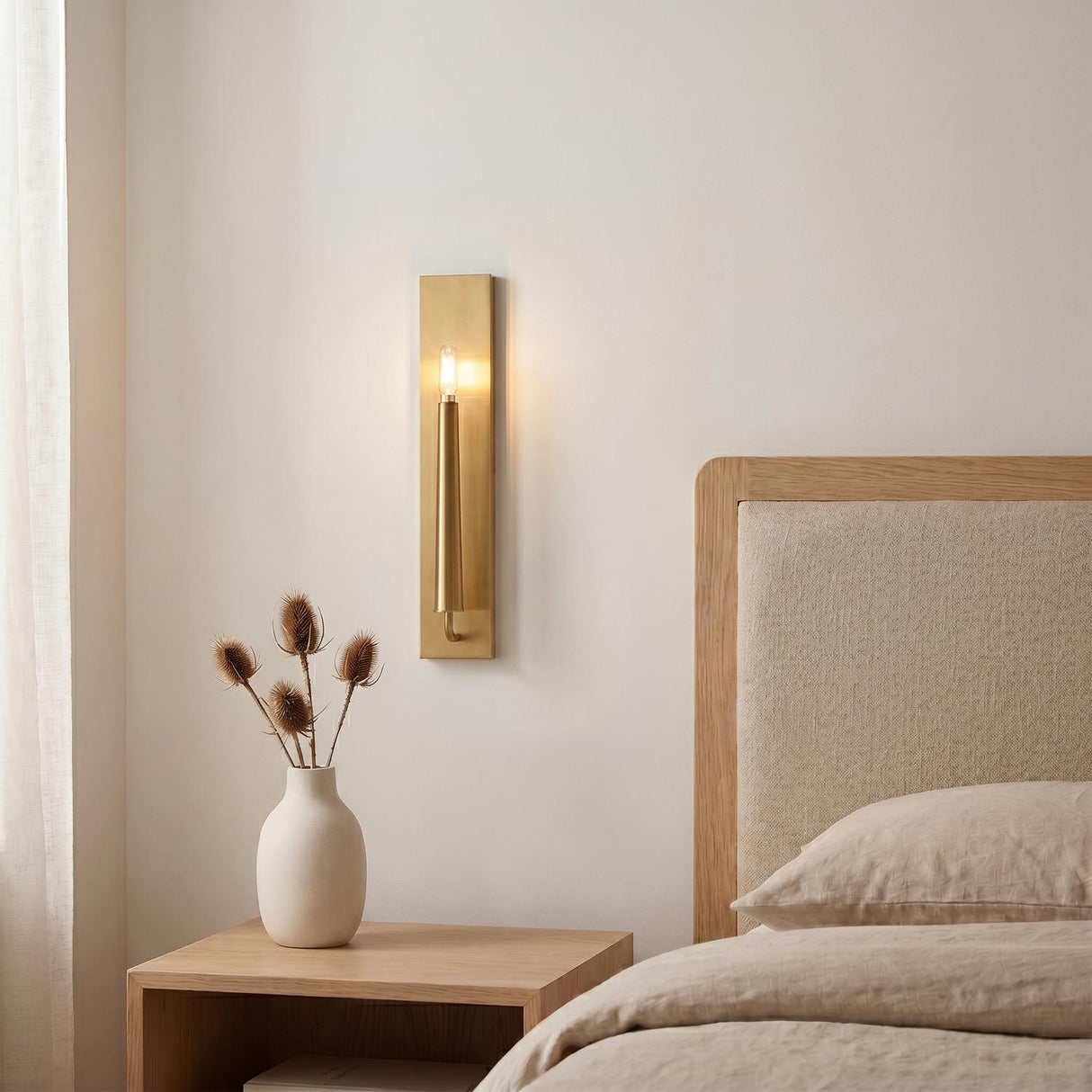 A modern bedroom with the Solana Wall Sconce 18" in modern brass mounted beside a linen headboard and a white ceramic vase of thistles.