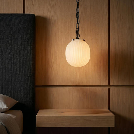 A mid-century bedroom with a matte black and ribbed milk glass globe pendant hanging over a floating wood nightstand beside a dark textured headboard.