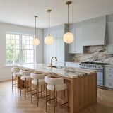 A transitional kitchen with three brushed brass and ribbed milk glass globe pendants hanging over a marble-topped wood island with light blue cabinetry and herringbone floors.