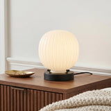 A mid-century bedroom featuring the Basile Small Table Lamp 8", Matte Black and Ribbed Milk Glass on a fluted walnut nightstand beside a cream knit blanket.