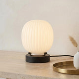 A modern living room featuring the Basile Small Table Lamp with matte black and ribbed milk glass on a travertine console table beside a round brass tray.