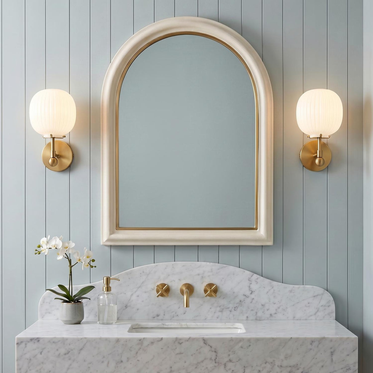 A transitional bathroom with two brushed brass and ribbed milk glass wall sconces flanking an arched mirror above a marble vanity with blue shiplap walls.