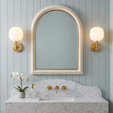 A transitional bathroom with two brushed brass and ribbed milk glass wall sconces flanking an arched mirror above a marble vanity with blue shiplap walls.