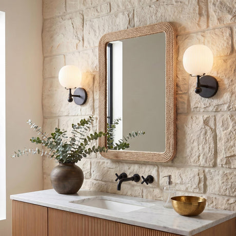 A coastal bathroom with the Basile Wall Sconce 12", Matte Black and Ribbed Milk Glass flanking a rope-wrapped mirror on a textured stone wall above a marble counter.